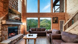 L’intérieur chaleureux d’un chalet avec une cheminée en pierre, des canapés en cuir et de grandes fenêtres donnant sur une colline boisée.