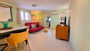 Ein Wohnzimmer mit einem roten Sofa, einem Esstisch mit Stühlen, einem kleinen runden Tisch und einem Fernseher auf einer Holzkommode.