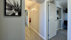 Un pasillo con un cuadro enmarcado de palmeras, un extintor y una puerta que da a una habitación con tocador y espejo.