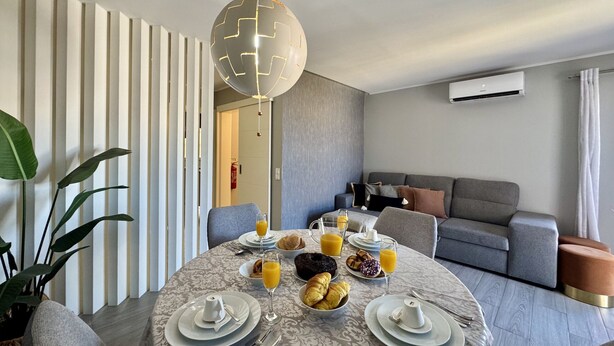 Un comedor moderno con una mesa redonda dispuesta para el desayuno, un sofá gris al fondo y una lámpara decorativa grande.