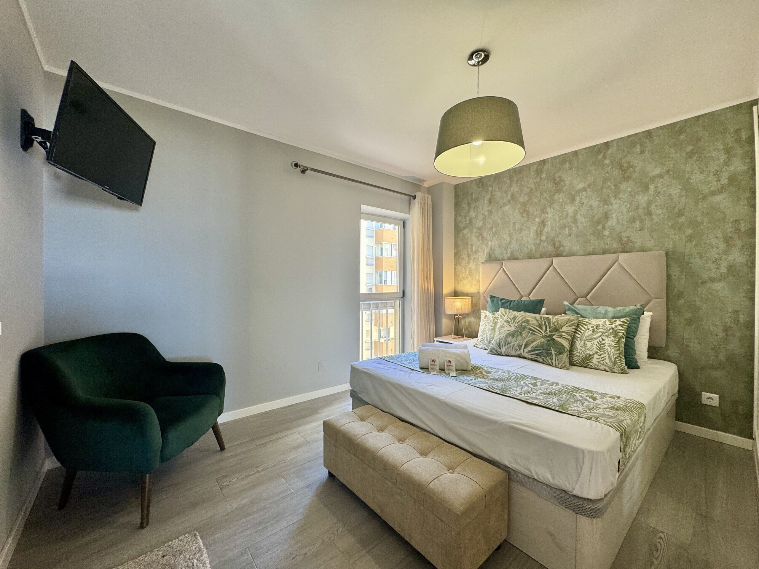 A bedroom with a bed, a chair, a television mounted on the wall, and a window.