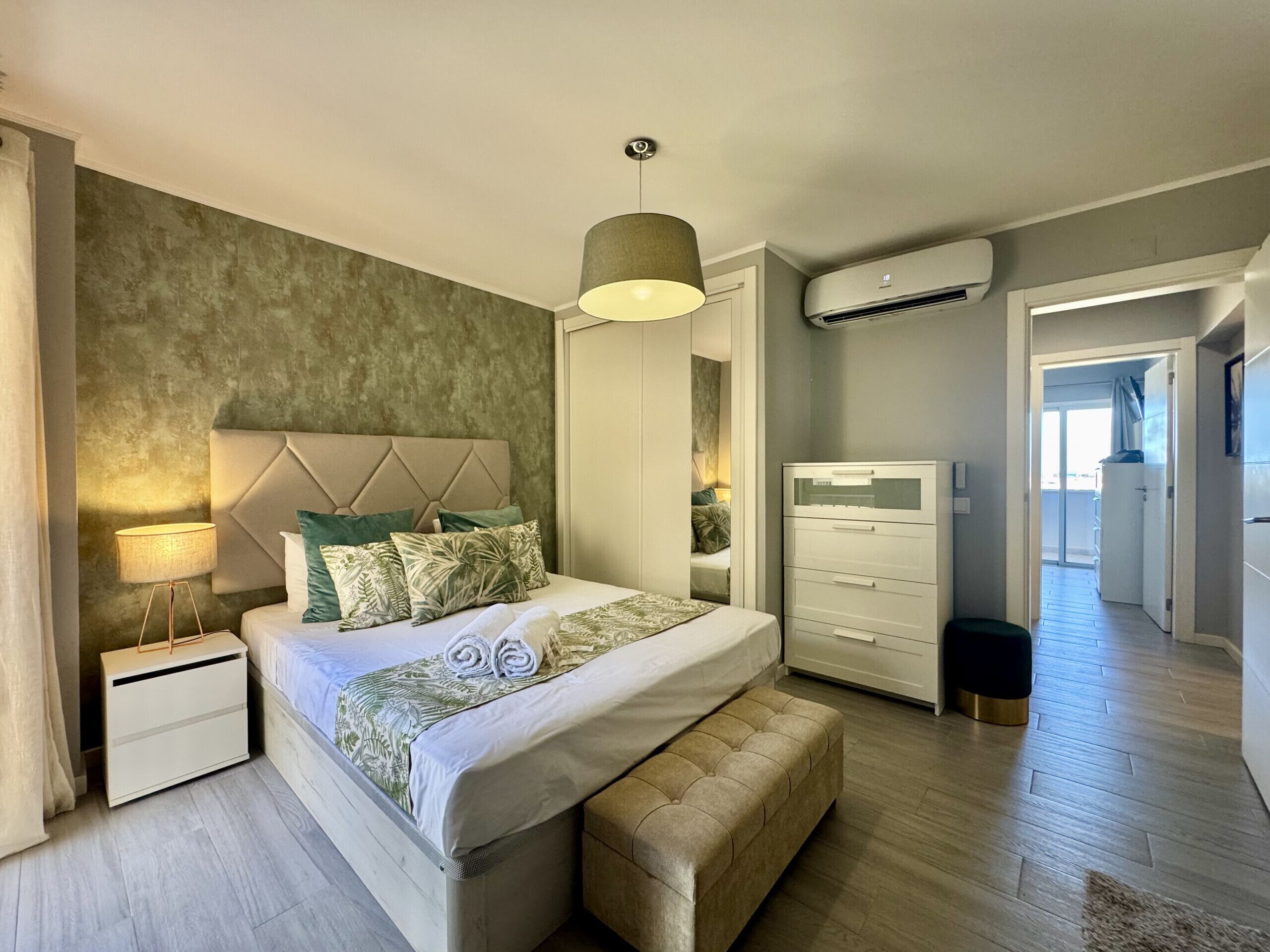 Une chambre à coucher comprenant un lit, des tables de chevet, une lampe, un appareil de climatisation, une armoire et donnant sur le couloir.