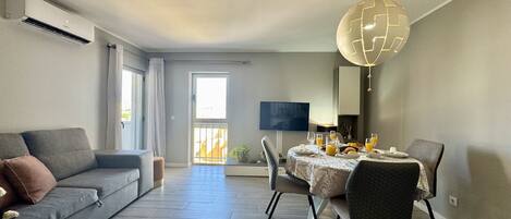 A modern living room with a grey sofa, a dining table set with breakfast, and a TV mounted on the wall.