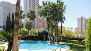 A swimming pool surrounded by palm trees and greenery, with tall buildings in the background.