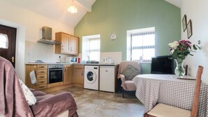 A compact kitchen with a washing machine, microwave, and stove. There is a chair with a blanket, a table with a vase, and a TV on a stand. - The Cow Byre (Saltburn-by-the-Sea)