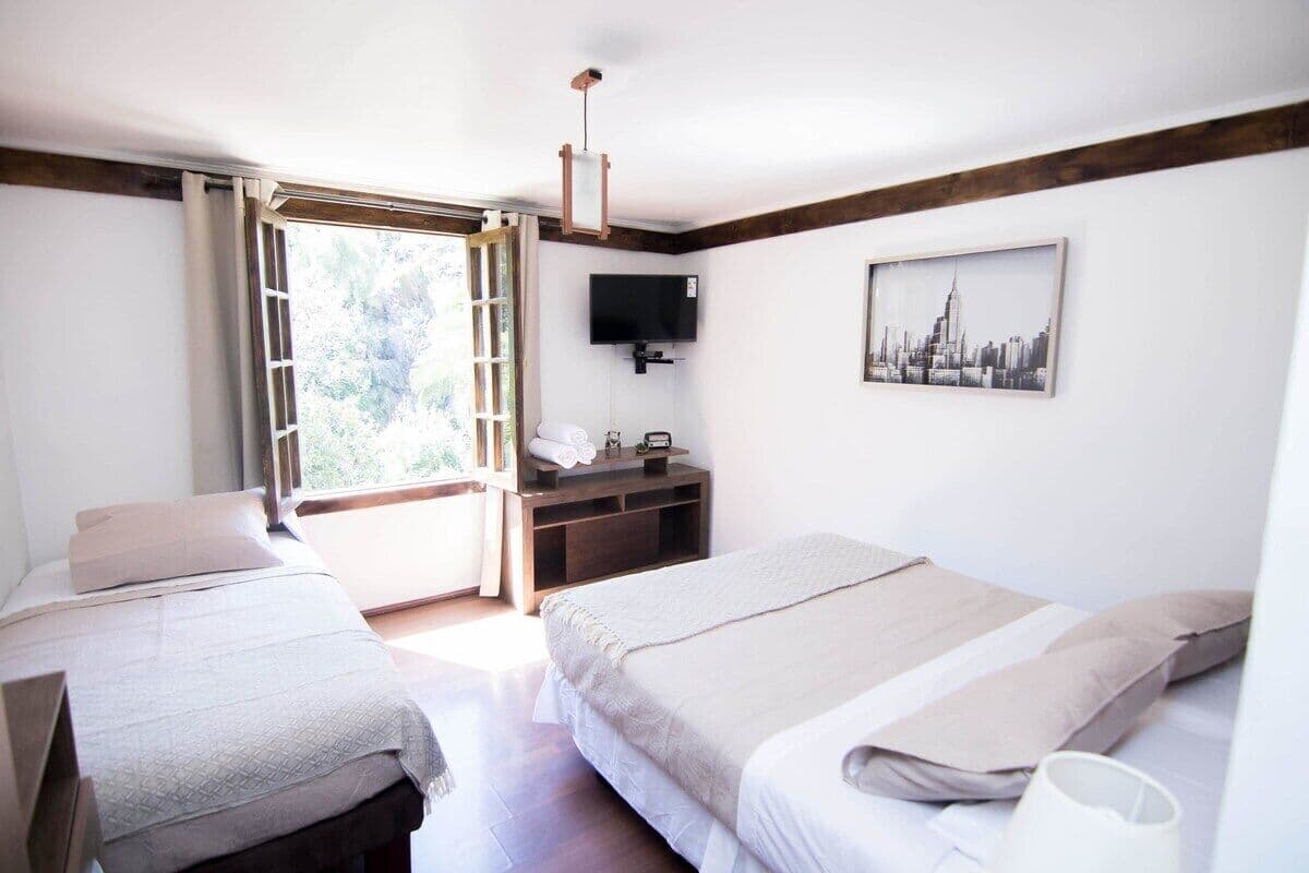 Un dormitorio con dos camas, un televisor, un cuadro con una vista urbana y una ventana con vistas a árboles.