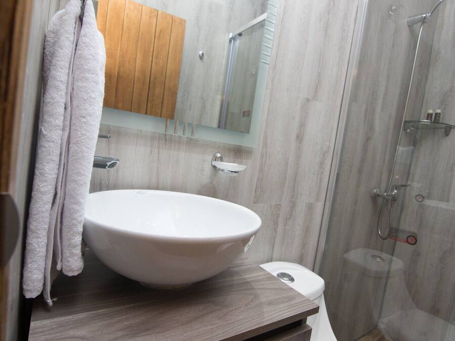 Un baño con lavamanos blanco, tocador de madera, cabina de ducha de vidrio y un mueble de madera sobre el espejo.