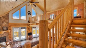 A spacious lodge with a stone fireplace, a wooden staircase, and a balcony with a view of the surrounding landscape. - Charming Log Cabin + Mountain Views + Fireplace + Hot Tub + Pet Friendly (Cripple Creek)