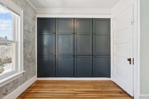 Ein Zimmer mit Holzboden, einem großen Fenster und einem dunkelbraunen Kleiderschrank mit mehreren Türen.