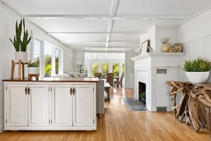Une cuisine spacieuse dotée d’un plancher en bois, de meubles blancs et d’un îlot central. On y trouve également un coin repas avec une table et des chaises, ainsi qu’une cheminée avec un manteau.