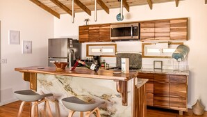 A modern kitchen with a marble island, wooden stools, a stainless steel refrigerator, and wooden cabinets.
