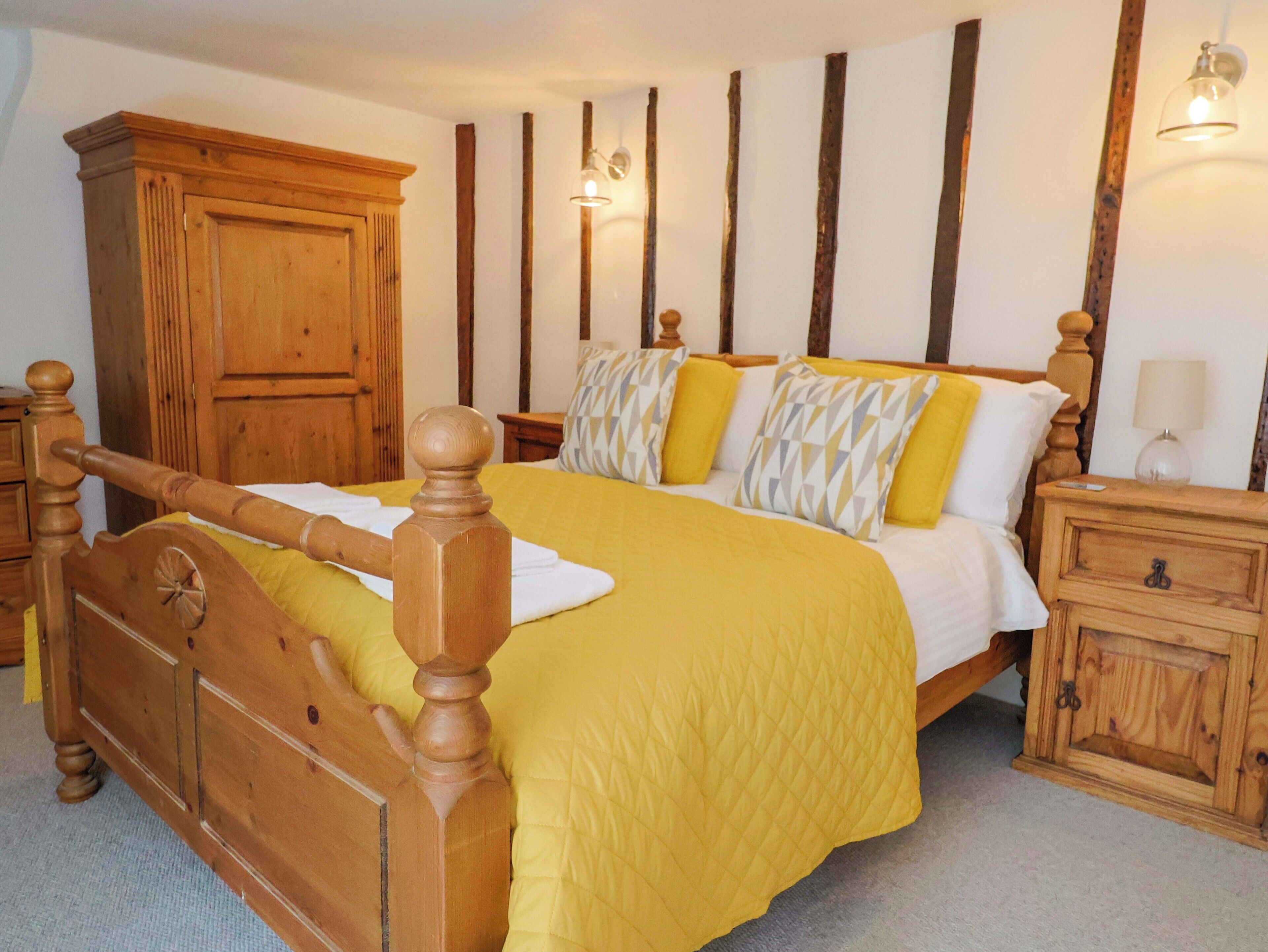 Ein hölzernes Himmelbett mit gelber Bettdecke, ein Nachttisch aus Holz mit einer Lampe und ein Holzschrank.