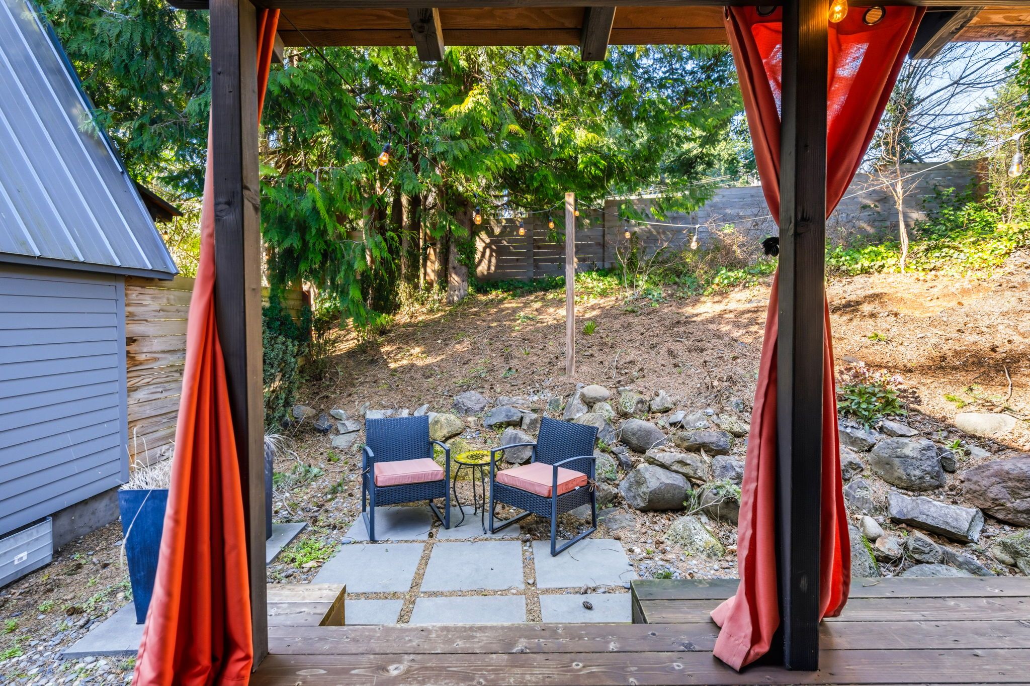 Un patio al aire libre con una pérgola de madera, dos sillas de mimbre con cojines, una mesita redonda y un sendero de piedra que lleva a una terraza de madera.
