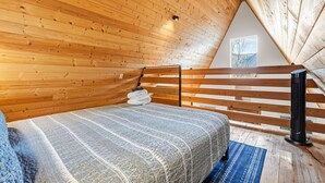 Een slaapkamer met een schuine kap, een bed met een geborduurde deken, een elektrische kachel en een raam met uitzicht op bomen.