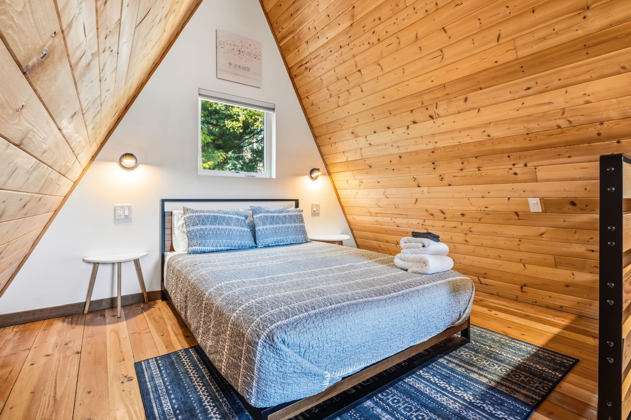 Un dormitorio con techo inclinado, una cama con colcha estampada, una mesita de noche pequeña, una lámpara fijada a la pared y un cuadro enmarcado sobre la cama.