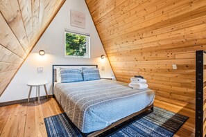 Un dormitorio con techo inclinado, una cama con colcha estampada, una mesita de noche pequeña, una lámpara fijada a la pared y un cuadro enmarcado sobre la cama.