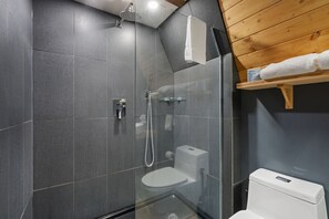 Un baño moderno con ducha de vidrio, inodoro blanco y estante de madera con toallas.
