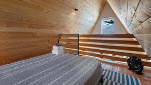 Quarto em estilo loft com teto inclinado, vigas de madeira, cama com colcha estampada, ventilador e vista para o exterior.