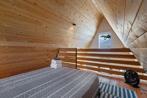 Un dormitorio tipo loft con techo inclinado, vigas de madera, cama con colcha estampada, un ventilador y vistas al exterior.