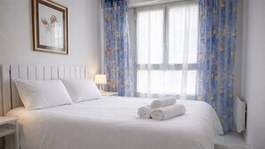 Una cama bien hecha con sábanas blancas y dos toallas enrolladas, una mesita de noche con lámpara y una obra de arte enmarcada en la pared.