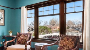 A room with two wooden chairs with floral cushions, a small round table, and a view of a residential area through large windows. - Historic Prescott Craftsman | Walk Downtown (Prescott)