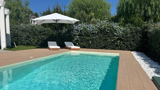 Una piscina rettangolare con acqua cristallina, circondata da un solarium in legno. Sono presenti due sdraio bianche e un ombrellone bianco che offre ombra.