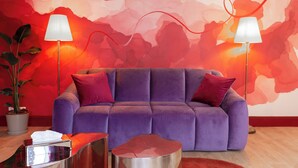 Ein violettes Sofa mit roten und weißen Kissen, zwei Stehlampen, ein Topfpflanze und ein metallischer Beistelltisch mit einer Taschentuchbox.