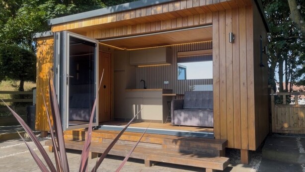 Eine moderne Holz-Blockhütte mit offener Tür, durch die eine Küche und ein Wohnbereich Einblick gewähren.