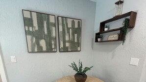 Un comedor con una mesa redonda de madera, cuatro sillas, dos cuadros abstractos colgados en la pared, una planta en maceta y un estante con adornos.
