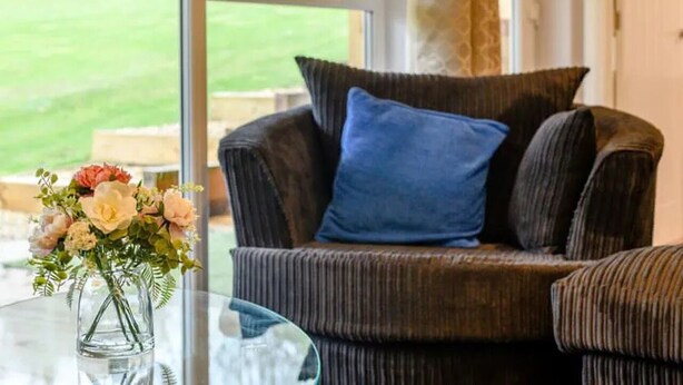 Ein dunkelbraunes Polstersofa mit einem blauen Kissen, platziert neben einem Glastisch mit Blumen in einer Vase.