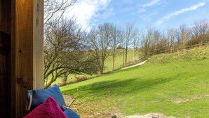 Eine hölzerne Bank mit Kissen, ein Glas Wasser und ein weiteres Glas Wasser auf einer Ablage mit Blick auf ein Wiesfeld und Bäume.