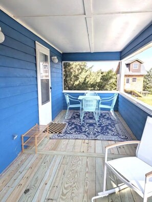 Un porche cubierto con fachada azul, piso de madera, una mesa con sillas y vista a una casa al fondo.