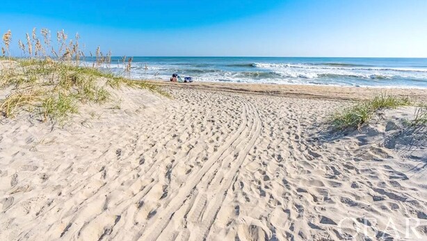 A sandy beach with footprints leading to the water, a dune with grass, and a vehicle parked near the water.