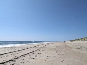 A beach with footprints leading to the water, a clear sky, and distant dunes.