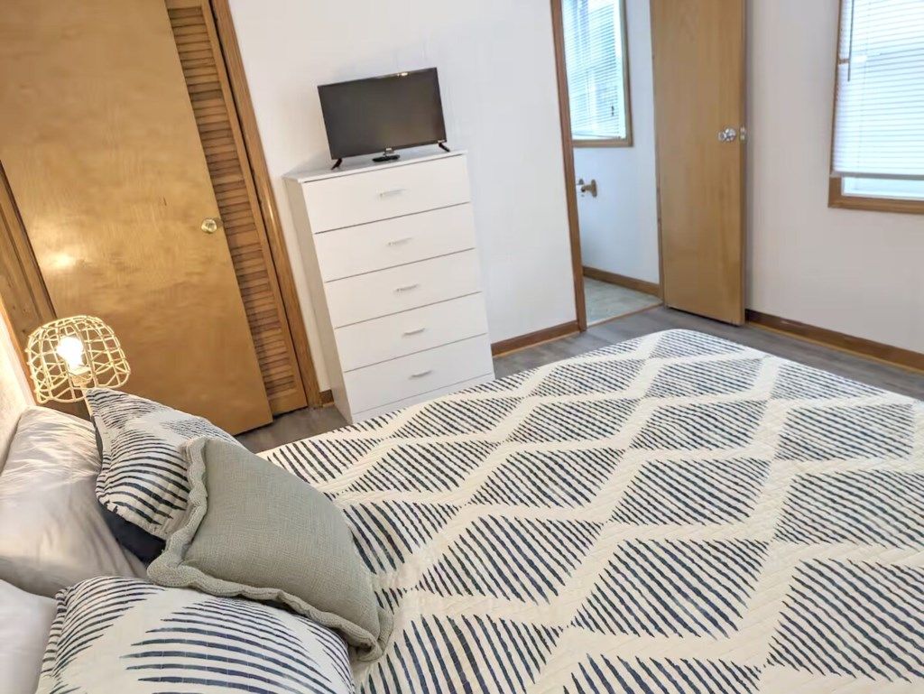 A bedroom with a bed, a dresser, a television on top, and a window with blinds.