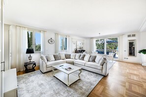 Ein geräumiges Wohnzimmer mit einer großen, L-förmigen Eckcouch, einem Couchtisch und Blick ins Freie durch große Fenster.