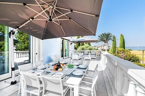 Un área de comedor al aire libre con muebles blancos, un paraguas grande y vista al mar.
