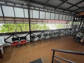 A covered outdoor seating area with a wooden floor, metal railings, and a view of greenery outside. - Slinky Hostel (Ko Phi Phi)