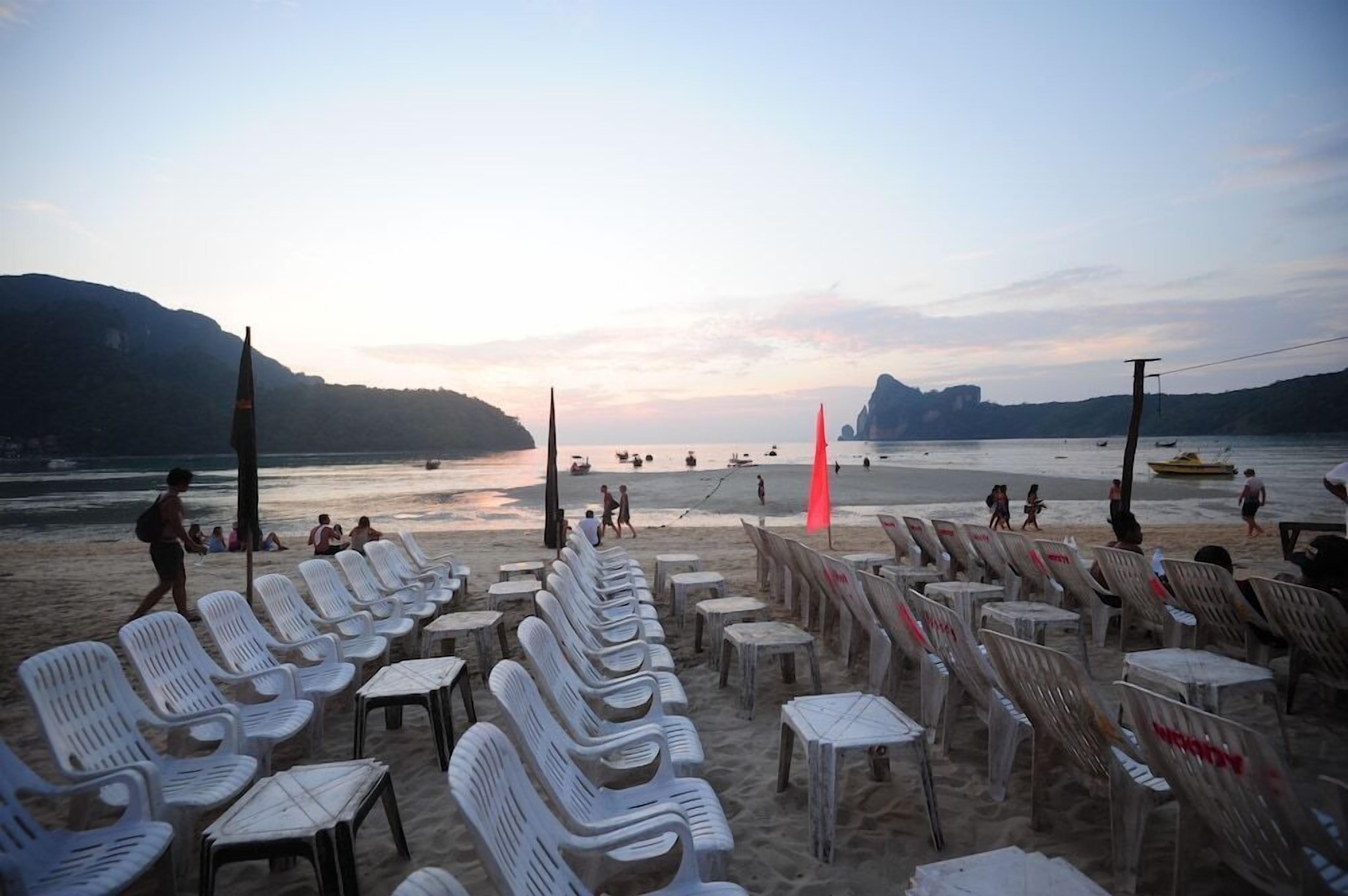 A beach with rows of white plastic chairs and tables, a few people walking, and boats on the water.