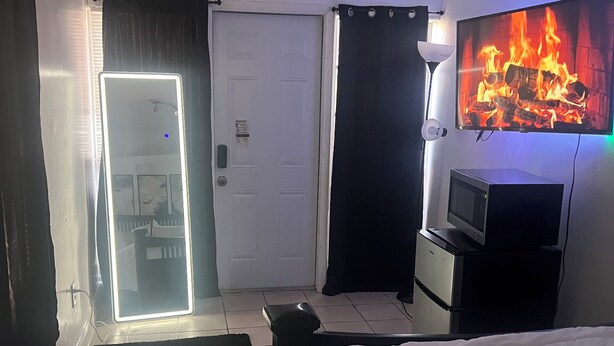A hotel room with a bed, a TV mounted on the wall, a door with a light, and a small desk with a lamp. - Cozy Studio in Miramar (Miramar)