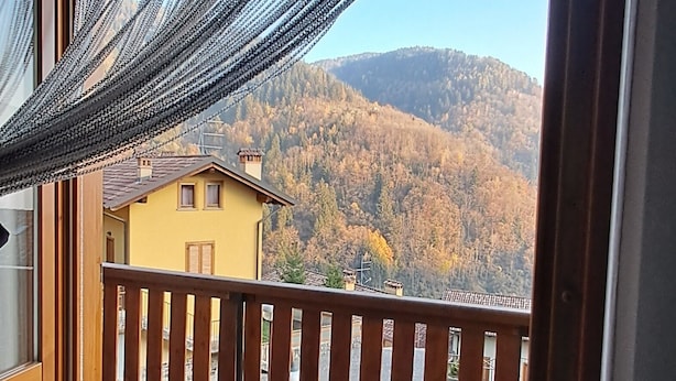 A balcony with a view of a yellow building and a forested mountain. - House of the stars (Lombardia)