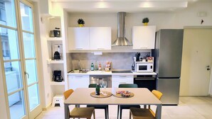 A modern kitchen with white cabinets, a stainless steel range hood, and a built-in microwave. There is a dining table with two chairs and a bottle of champagne.
