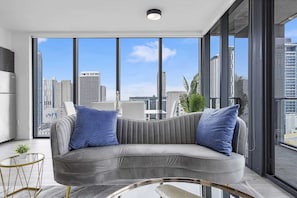 A modern living room with a grey sofa, two blue pillows, a glass coffee table, and a view of a cityscape through large windows. - X Lounge VII 3BR/3 Ensuite/Private Sky Residence (Miami)