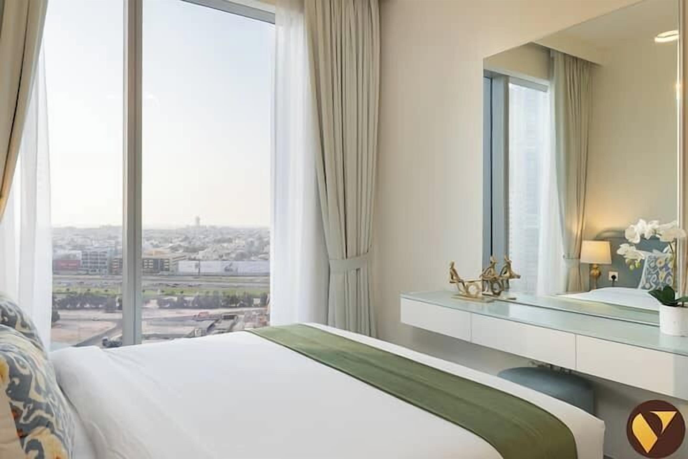 A hotel room with a large window, a bed with a green bedspread, a vanity with a mirror, and a view of the cityscape.