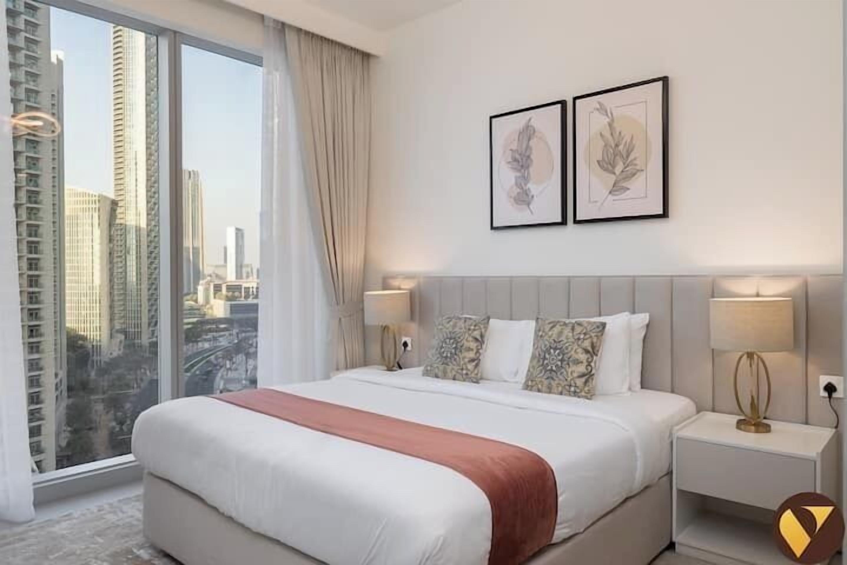 A hotel room with a large bed, two bedside lamps, a view of tall buildings, and two framed artworks on the wall.