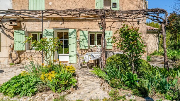 Un bâtiment rustique en pierre, doté de volets verts, un jardin avec des fleurs jaunes et une table avec des chaises à l’extérieur.