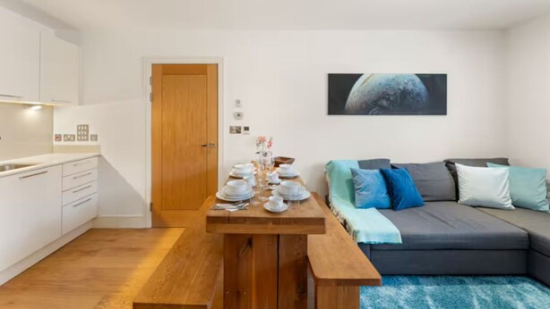 Een moderne woonruimte met een houten eettafel gedekt voor vier personen, een grijze bank met blauwe kussens, en een keuken met witte kastjes.