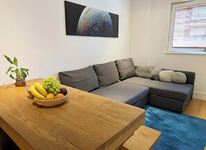 Un salon moderne avec un canapé d’angle, une table basse en bois sur laquelle est posé un panier de fruits, une plante en pot et une grande œuvre d’art accrochée au mur.