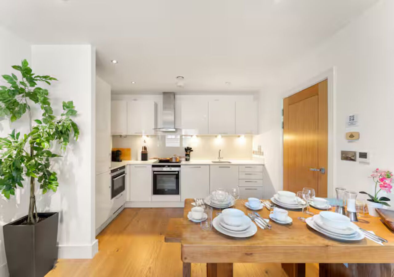 A modern kitchen with white cabinets, a wooden dining table set for four, and a potted plant.