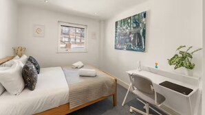 Een slaapkamer met een bed, een bureau, een stoel, een raam en een schilderij aan de muur.